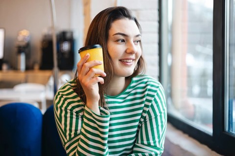 Woman smiling and holding a coffee cup, sitting by the window in a cafe, wearing a striped shirt