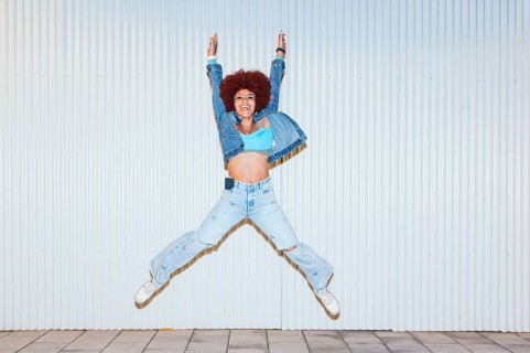 Exuberant African descendent woman with curly afro hair, jumping energetically in ripped jeans and