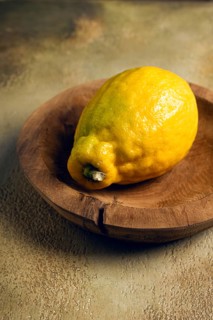 A vibrant yellow lemon sits in a hand-carved wooden bowl. The lemon\'s textured skin shines in