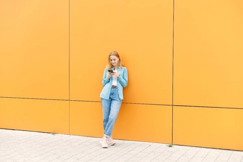 A woman in casual attire leans against a vibrant yellow wall, engaging with her smartphone The