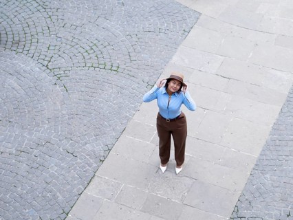 A high-angle view of a woman dressed in a blue shirt and brown pants, adjusting her hat on a