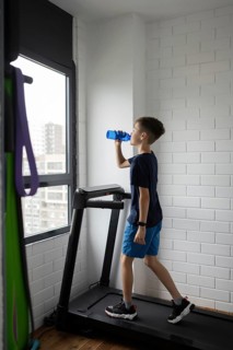 A boy in blue shorts and a dark shirt drinks water while on a treadmill next to a bright window. He