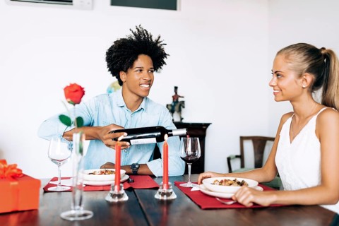 A man and woman share a romantic dinner at home, featuring wine, red candles, and a rose. The