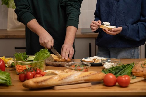 People preparing a fresh sandwich with a crusty baguette, cream cheese, and various vegetables.