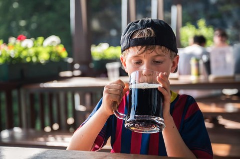 Cute boy is drinking a black drink from a large mug at a restaurant outdoor table