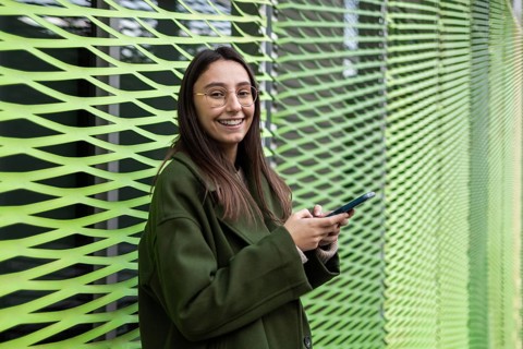 A woman wearing a green coat smiles as she uses her smartphone outside The lively green backdrop