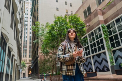 Young woman navigating urban streets, looking up with curiosity while holding her smartphone,