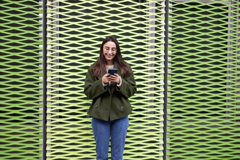 A woman with glasses and a green jacket stands smiling, using her phone against a unique green