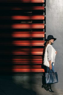 A fashionable woman in a wide-brimmed hat stands against a striking red wall. The contrast of light