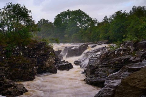 A bubbling river flows through a tree-lined, rocky landscape under cloudy skies, River Orchy, Eas