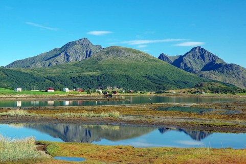 A calm body of water reflects mountains and a small village with clear skies, Leknes, Vestvågøya,