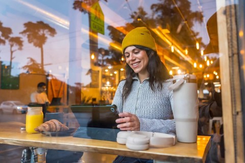 Young woman wearing a yellow beanie managing work on a digital tablet in a coffee shop, enjoying a