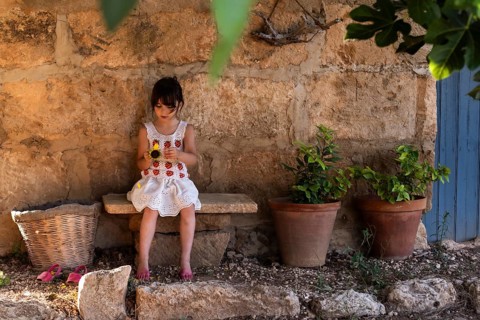 A young girl in a white dress sits on a stone bench, holding a sunflower. She is surrounded by