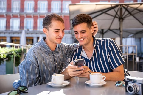 Smiling gay couple enjoying coffee while browsing a smartphone at an outdoor cafe in Plaza Mayor,
