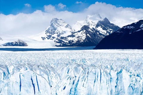 Perito Moreno glacier with rugged ice surface, snow-capped Andean mountain peaks under cloudy sky,