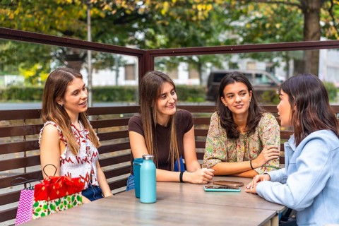 Four young women chatting and laughing at an outdoor cafe table after a shopping spree, enjoying