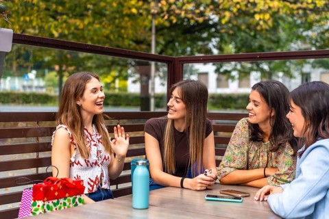 Four young women friends enjoying conversation and shared laughter at a cafe table outdoors,