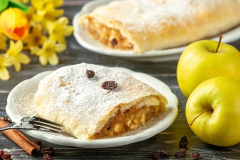 Apple strudel on a plate with powdered sugar and raisins, yellow apples and spring flowers