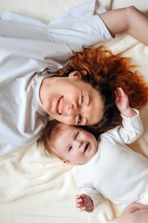 A joyful mother and her adorable baby girl lying together on a cozy bed, both dressed in white. The