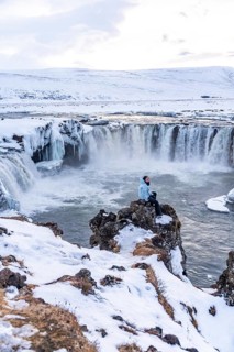 Female hiker sitting on frozen and snowy Godafoss waterfall in winter in Iceland