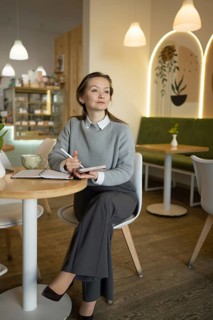 A woman in a stylish outfit sits thoughtfully in a cozy cafe, holding a pen and notebook. The warm