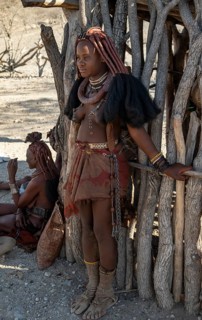 Young traditionally dressed Himba woman, leaning against a wooden hut, near Opuwo, Kaokoveld,