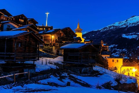 Snow-covered roofs of old farmhouses and barn storage, in the back the illuminated cross and the