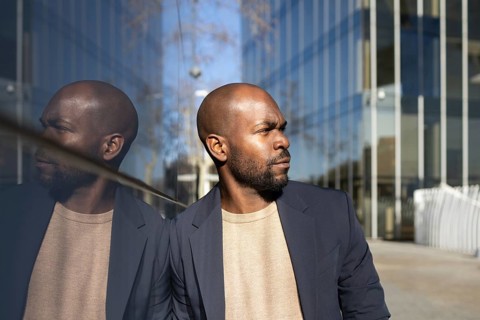 Confident cuban man in a blazer stands near a reflective glass wall, his reflection visible. Modern