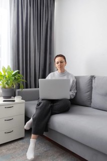 A woman wearing glasses works on a laptop while sitting on a gray sofa in a bright room. A potted