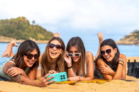 Four happy young women lying on a sandy beach, wearing sunglasses, laughing and taking a selfie
