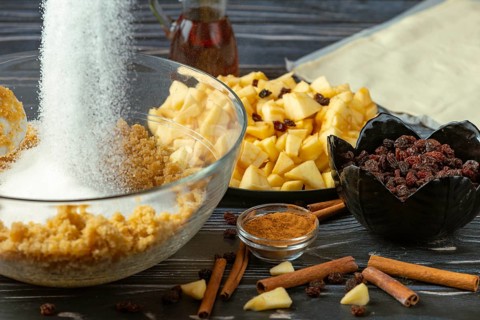 Sugar is poured into a bowl with breadcrumbs, next to apple pieces, raisins and cinnamon
