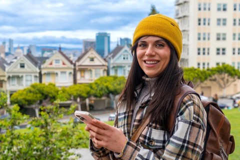 Young woman smiling at the camera, holding a smartphone, with the iconic colorful victorian houses