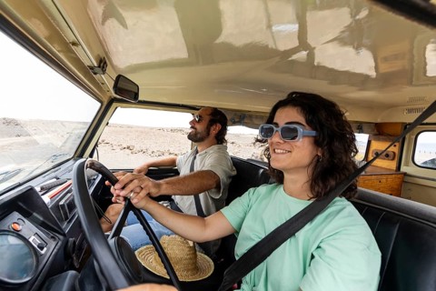 A blind man sits in the passenger seat while a woman drives a motorhome through a scenic desert.