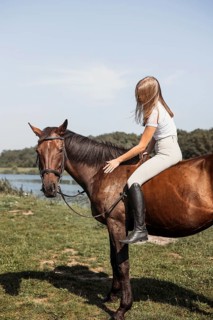 A young girl rides a brown horse near a serene lake on a sunny day. She wears casual riding attire,
