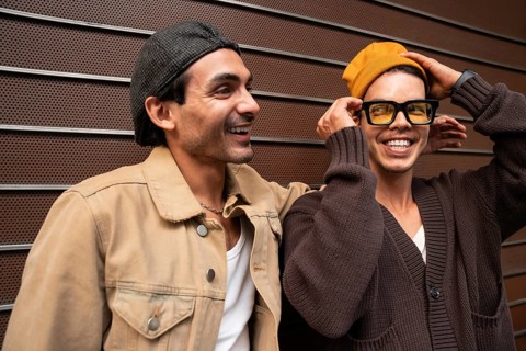 A cheerful couple stands side by side against a modern metal wall. Both are dressed in casual urban