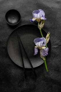 Top view of elegant iris flowers alongside black ceramic dishware on a textured dark background,