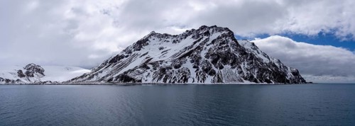 Snow-capped mountain with rugged rock formations rising above the ocean, cloudy sky, coastal