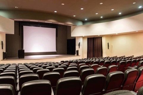 An empty convention center with a large white screen and rows of seats. The stage is set for an
