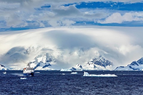 A ship sails in the ocean, surrounded by icebergs and snowy mountains under a cloudy sky, cruise