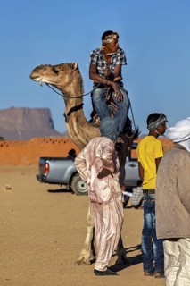 A man on a camel stands next to people in traditional dress against a mountain backdrop, Tuareg
