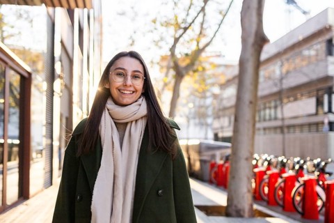 A cheerful woman in a green coat smiles warmly on a sunny day in an urban environment She is
