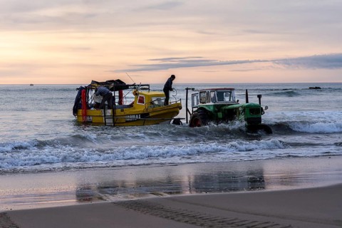 Yellow fishing boat with two fishermen is pulled from beach into water by green tractor at sunrise,