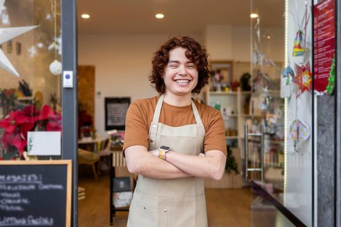 A cheerful young shop assistant in an apron stands with crossed arms at the entrance of an organic
