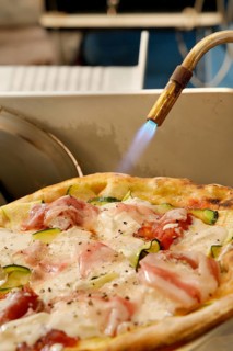 Blowtorch being applied to a gourmet pizza to crisp up toppings. The flame gently caramelizes the