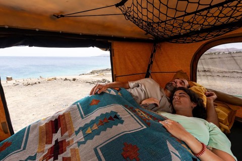 A blind man rests in a motorhome with his partner, wrapped in a colorful blanket, gazing out at the