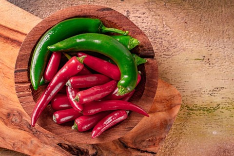 Bright green and red peppers are neatly placed in a round wooden bowl on a wooden board. The board