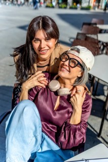 Two womenone younger, one olderembrace and smile at the camera while sitting at an outdoor cafe,
