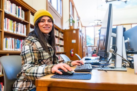 Young woman wearing a beanie and plaid shirt smiling at the viewer while researching and typing on