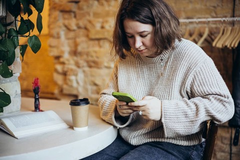 A young woman in a relaxed sweater sits in a cozy bookstore cafe, looking at her smartphone A