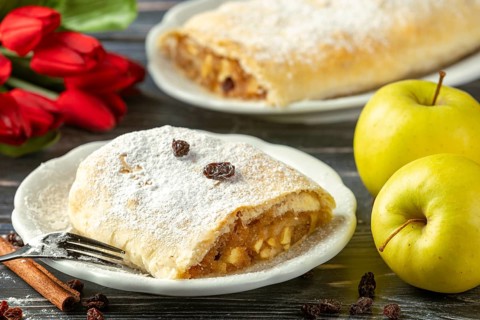 Apple strudel on a plate with powdered sugar and raisins, next to yellow apples and red flowers